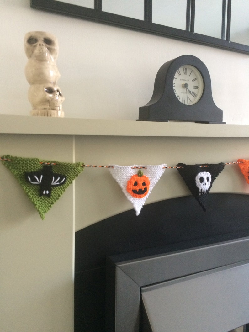May include: A knitted Halloween banner with green, white, orange, and black triangular flags. Each flag features a different Halloween icon: a bat, a pumpkin, a skull, and a ghost. The banner is hanging above a fireplace.