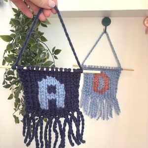 Op de afbeelding: Twee gebreide wandkleden. Eén is marineblauw met een lichtblauwe letter "A" en franjes. De andere is lichtblauw met een koraalkleurige letter "D". Beide zijn opgehangen met een marineblauw koord en een houten stok.