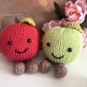 Peut inclure: Deux jouets en forme de pomme tricotés, un rouge et un vert. Chacun a des yeux noirs en forme de bouton, un sourire cousu, une tige marron et une feuille verte. Ils ont de petits pieds marron tricotés. Le fond comprend des fleurs roses.
