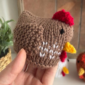May include: A handmade brown knitted chicken with a red comb and yellow beak. The chicken has white accents on its body and small black eyes. The image shows a close-up of the chicken, with other similar knitted chickens in the background.