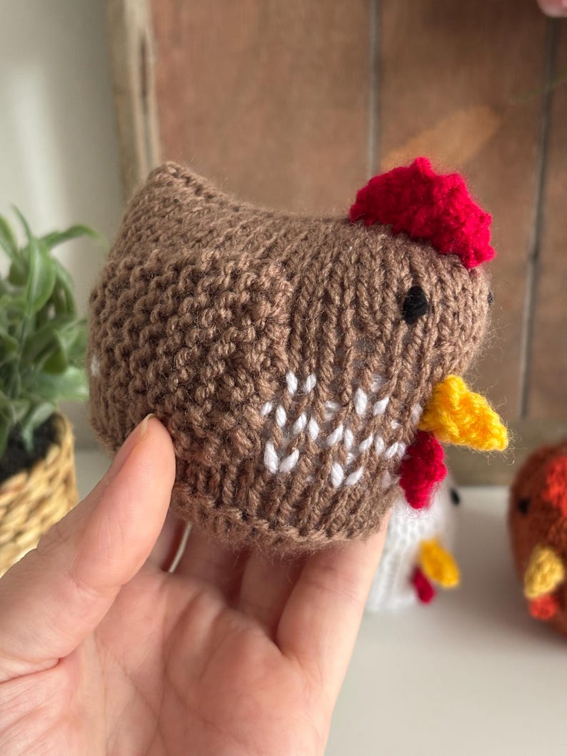 May include: A handmade brown knitted chicken with a red comb and yellow beak. The chicken has white accents on its body and small black eyes. The image shows a close-up of the chicken, with other similar knitted chickens in the background.