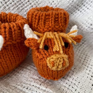 May include: A pair of knitted baby booties with a brown and white highland cow design. The booties are made with a soft, textured yarn and have a cute, whimsical look.