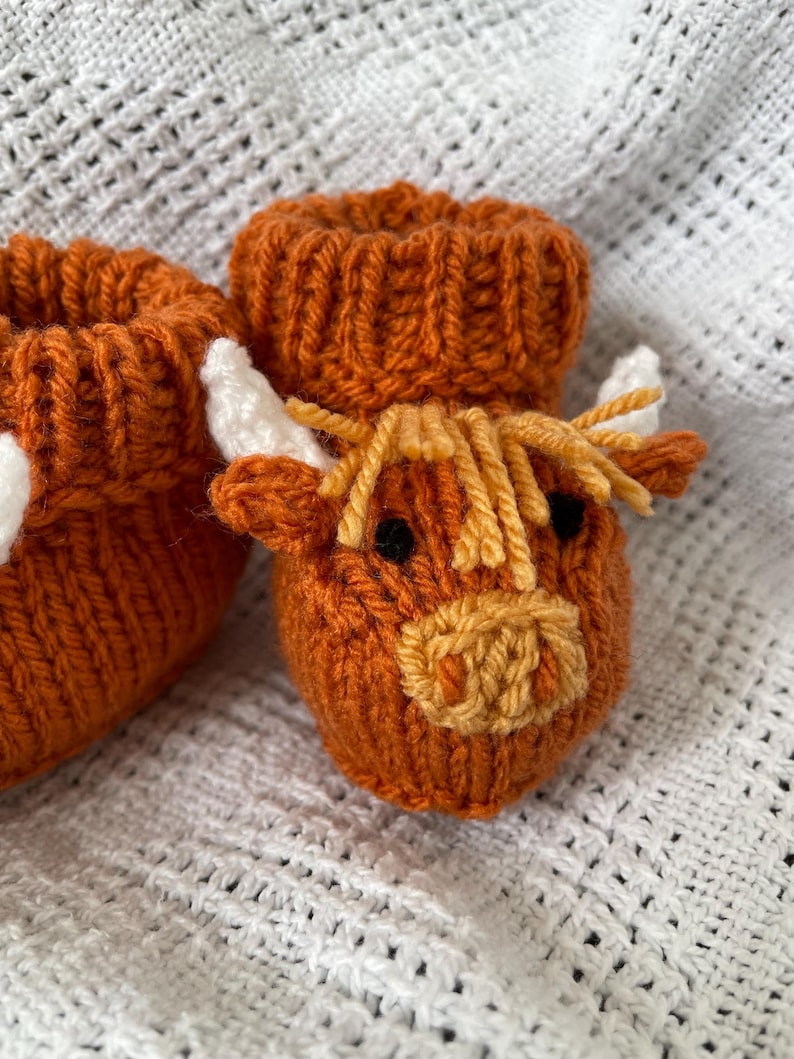 May include: A pair of knitted baby booties with a brown and white highland cow design. The booties are made with a soft, textured yarn and have a cute, whimsical look.