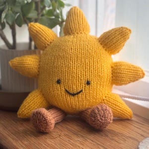 Op de afbeelding: Een handgemaakt, gebreid zonvormig pluche speelgoed. De zon is voornamelijk geel met bruine voetjes. Het heeft een lachend gezicht met zwarte ogen en een gebogen mond. De zonnestralen zijn puntig en strekken zich uit.