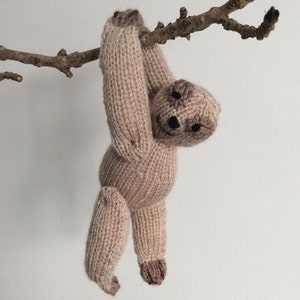 May include: A knitted sloth toy hanging from a branch. The sloth is brown and has a cute face.