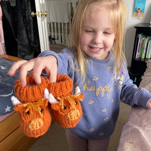 May include: A young girl is holding up two orange knitted cow-shaped baby booties. The booties have white horns and black eyes. The girl is wearing a purple shirt with a pattern of bees and the word "happy" on it.