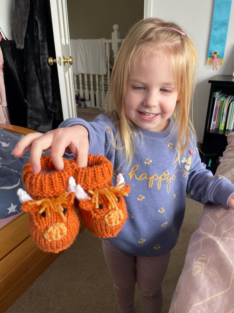 May include: A young girl is holding up two orange knitted cow-shaped baby booties. The booties have white horns and black eyes. The girl is wearing a purple shirt with a pattern of bees and the word "happy" on it.