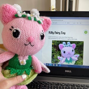 May include: A pink knitted fairy toy with green wings and a white flower crown. The toy is wearing a green dress with a white flower on the front. The toy is sitting on a laptop with a knitting pattern for a similar toy on the screen. The pattern is titled "Kitty Fairy Toy".