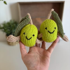 Pode incluir: Dois brinquedos de pelúcia de azeitona verde tricotados com rostos sorridentes e folhas presas. As azeitonas são seguradas em uma mão, mostrando os detalhes feitos à mão e o design alegre. Os brinquedos de pelúcia têm caules castanhos e folhas verdes.