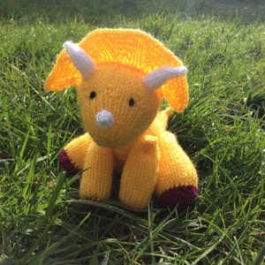 May include: A yellow knitted triceratops toy with white horns and a burgundy nose. The toy is sitting on a bed of green grass.