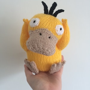 May include: A yellow knitted plush toy with a brown beak and black spiky hair. The toy has white eyes and is holding its arms up.