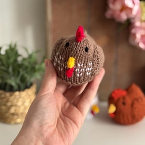 May include: A brown knitted chicken with a red comb and yellow wattle. The chicken has black eyes and white and brown patterned details. Another similar chicken is visible in the background. The chicken is held in a hand.