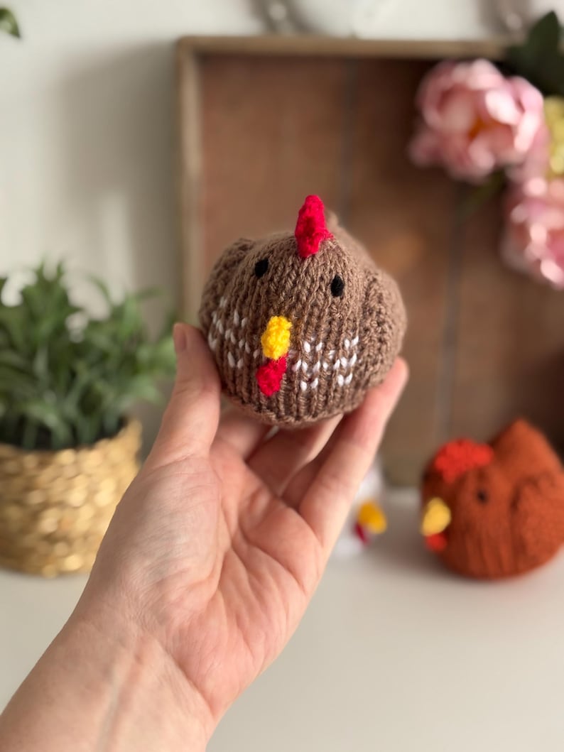 May include: A brown knitted chicken with a red comb and yellow wattle. The chicken has black eyes and white and brown patterned details. Another similar chicken is visible in the background. The chicken is held in a hand.
