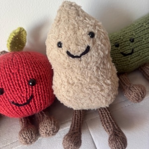 Op de afbeelding: Drie gebreide pluche speelgoed: een rode appel met een groen blad, een beige peer en een groene komkommer. Elk heeft een lachend gezicht en bruine gebreide benen. Het speelgoed is ontworpen met een zacht, getextureerd uiterlijk.