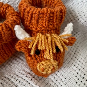 May include: A pair of knitted baby booties shaped like a Highland cow. The booties are brown with a yellow and white face and white horns.