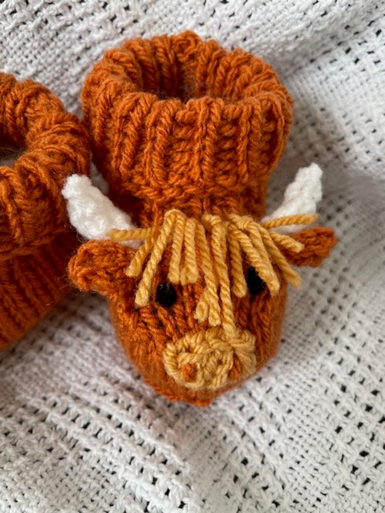 May include: A pair of knitted baby booties shaped like a Highland cow. The booties are brown with a yellow and white face and white horns.