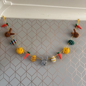 Op de afbeelding: Een handgemaakte paasslinger met gebreide konijntjes, kuikens, wortels en versierde eieren. De slinger is geregen aan een gele draad en hangt tegen een muur met geometrisch patroon. De kleuren zijn bruin, geel, oranje, groen en grijs.