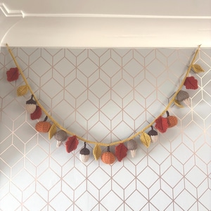 May include: A crocheted autumn garland with leaves, acorns, and pumpkins in red, orange, yellow, and brown. The garland hangs against a geometric patterned wall, providing a seasonal decorative accent.