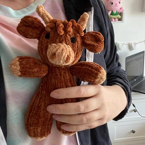 May include: A brown plush toy cow with a long, curly brown mane and a cream-colored face. The toy is being held by a person.