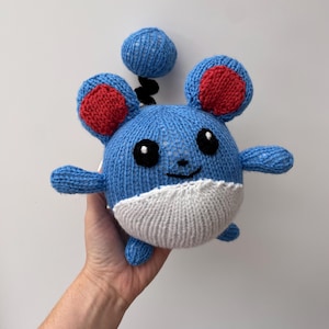May include: A blue and white knitted plush toy resembling a cartoon mouse with red ears and black eyes. The toy has a white belly and a black tail.