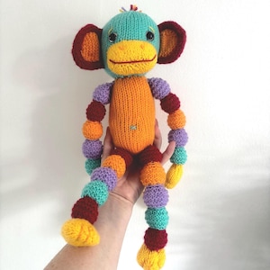 Puede incluir: Una muñeca de mono tejida a mano y colorida. El mono tiene una cabeza turquesa, un cuerpo naranja y extremidades articuladas con esferas de varios colores. Las orejas son de color rojo oscuro y la boca es amarilla. Es un juguete blando.