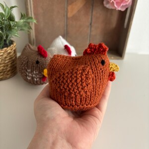 May include: A knitted chicken figurine in a warm orange hue, featuring a red comb and yellow wattle. The chicken is held in a hand, with two other chicken figurines visible. These charming, handcrafted chickens are a delightful item.