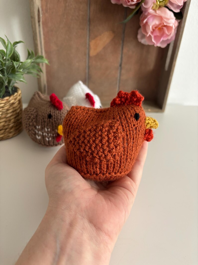May include: A knitted chicken figurine in a warm orange hue, featuring a red comb and yellow wattle. The chicken is held in a hand, with two other chicken figurines visible. These charming, handcrafted chickens are a delightful item.