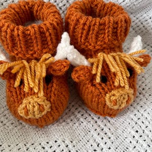May include: A pair of knitted baby booties shaped like brown Highland cows with white horns and yellow fur.