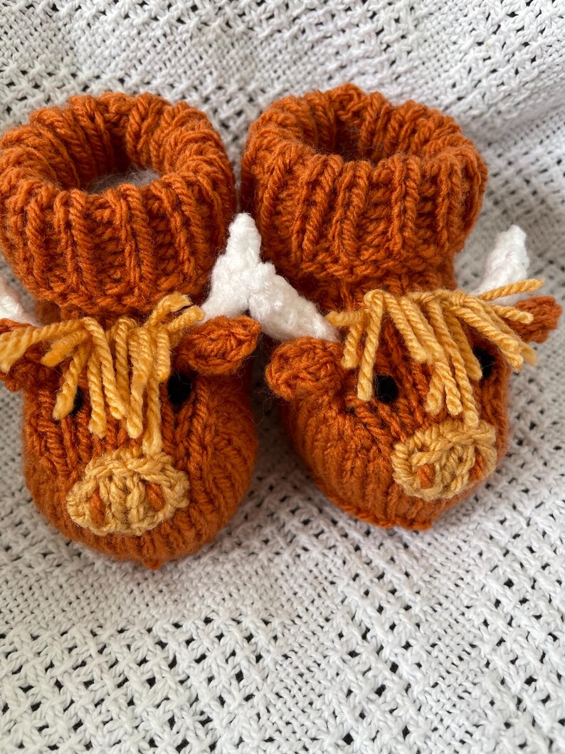 May include: A pair of knitted baby booties shaped like brown Highland cows with white horns and yellow fur.