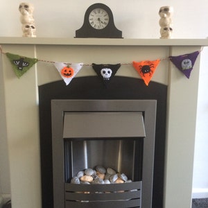 May include: A fireplace mantel decorated with a Halloween-themed knitted banner featuring a bat, pumpkin, skull, spider, and tombstone. The banner is strung with orange yarn and hangs above a black fireplace with a silver metal insert and a bed of gray and orange stones.