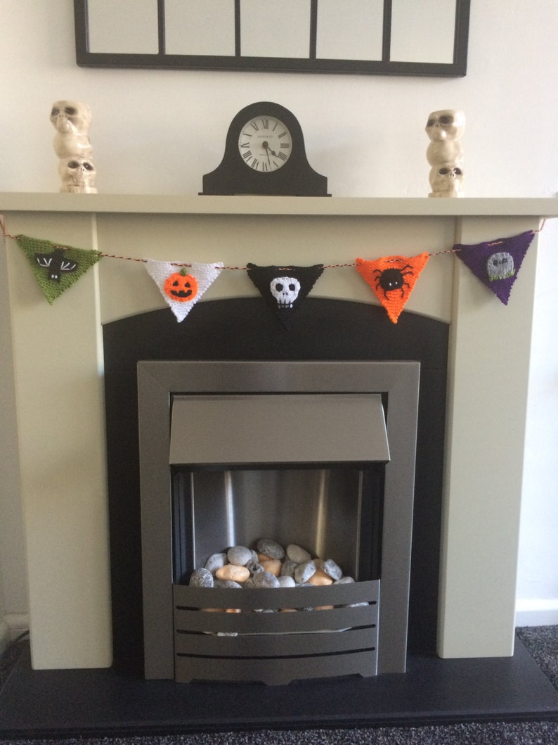 May include: A fireplace mantel decorated with a Halloween-themed knitted banner featuring a bat, pumpkin, skull, spider, and tombstone. The banner is strung with orange yarn and hangs above a black fireplace with a silver metal insert and a bed of gray and orange stones.