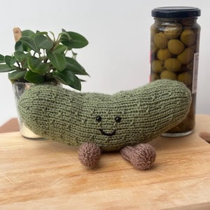 Peut inclure: Un jouet en peluche en forme de cornichon vert tricoté, avec un visage souriant et de petits pieds marron. Une petite plante en pot et un bocal d'olives sont en arrière-plan. La peluche est sur une planche à découper en bois.