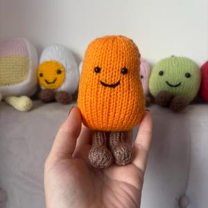 May include: A knitted orange pear-shaped plush toy with a smiling face and brown feet. Other knitted food-themed plush toys are visible in the background, including an egg and a green apple. The plush toy is held in a hand.