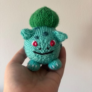 May include: A green knitted Bulbasaur plush toy with red eyes and a white smile. The toy is being held in a hand.