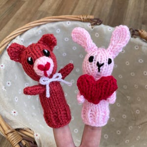 May include: Two finger puppets, one a red bear and the other a pink bunny, holding a red heart. The puppets are made of knitted yarn.
