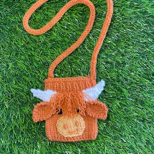 May include: A small orange knitted crossbody bag shaped like a Highland cow with white horns and a yellow smiling face.