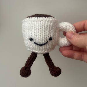 May include: A handmade, knitted coffee cup with a smiling face, white body, brown coffee, and brown legs. The cup has a white handle and black eyes. The coffee cup is a whimsical, handcrafted item.