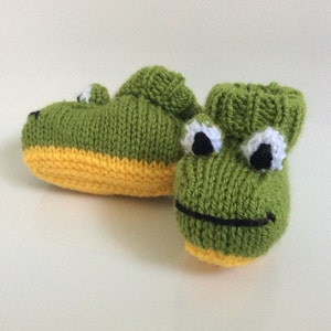 Könnte beinhalten: Ein Paar grüne gestrickte Babyschuhe mit gelben Sohlen und Froschgesichtern. Die Schuhe haben schwarze Augen, weiße Pupillen und schwarze Lächeln.