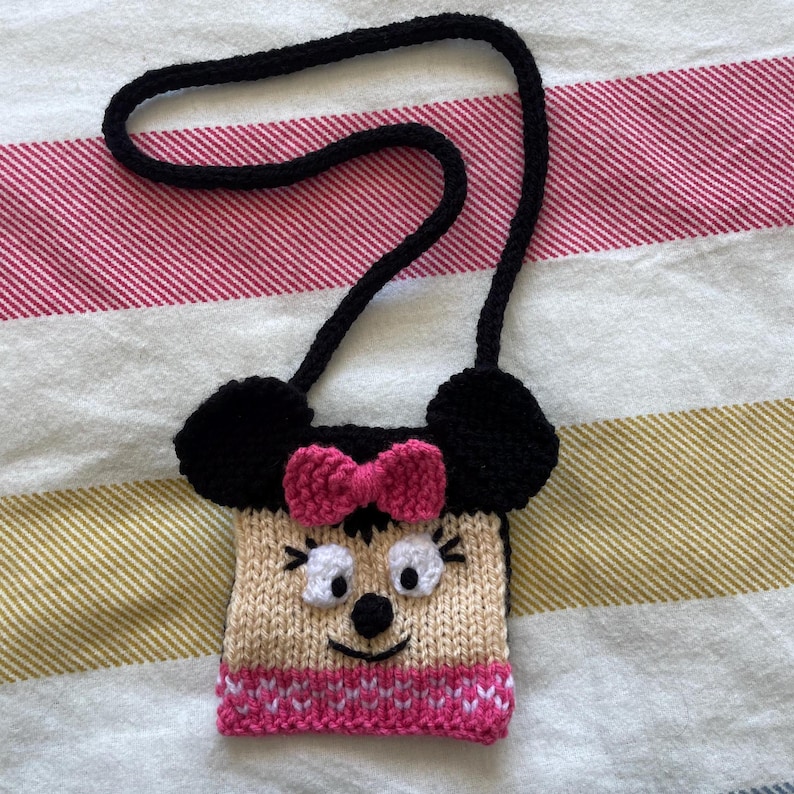Pu&ograve; includere: Una piccola borsa di maglia con una tracolla nera, a forma di topo dei cartoni animati con orecchie nere, un fiocco rosa e un fondo a righe bianco e rosa.