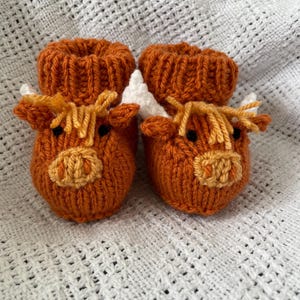 May include: A pair of knitted baby booties shaped like a brown Highland cow with white horns and yellow fur. The booties are on a white crocheted blanket.