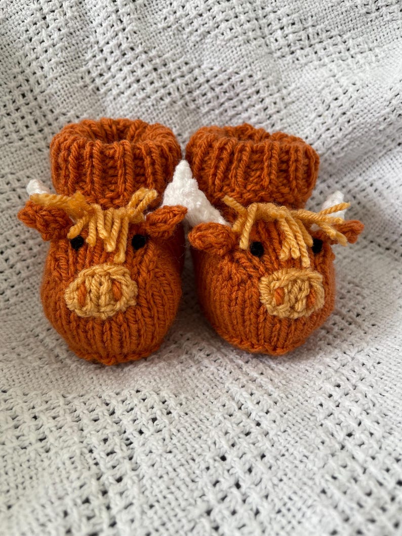 May include: A pair of knitted baby booties shaped like a brown Highland cow with white horns and yellow fur. The booties are on a white crocheted blanket.
