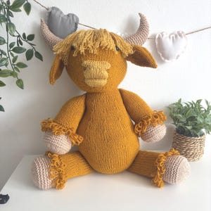 Op de afbeelding: Een handgemaakt, mosterdgeel gebreid Highland koeien knuffel met bruine hoorns en hoeven. De knuffel heeft een ruige manen en decoratieve afwerking op de hoeven en armen. Het speelgoed zit op een wit oppervlak.