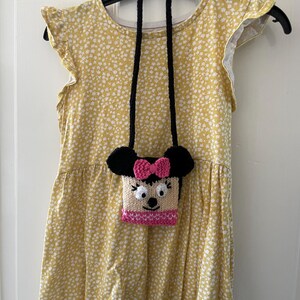 Pu&ograve; includere: Un vestito giallo con stampa floreale bianca e una borsa di maglia nera e rosa con un viso di cartone animato appeso alla tracolla.