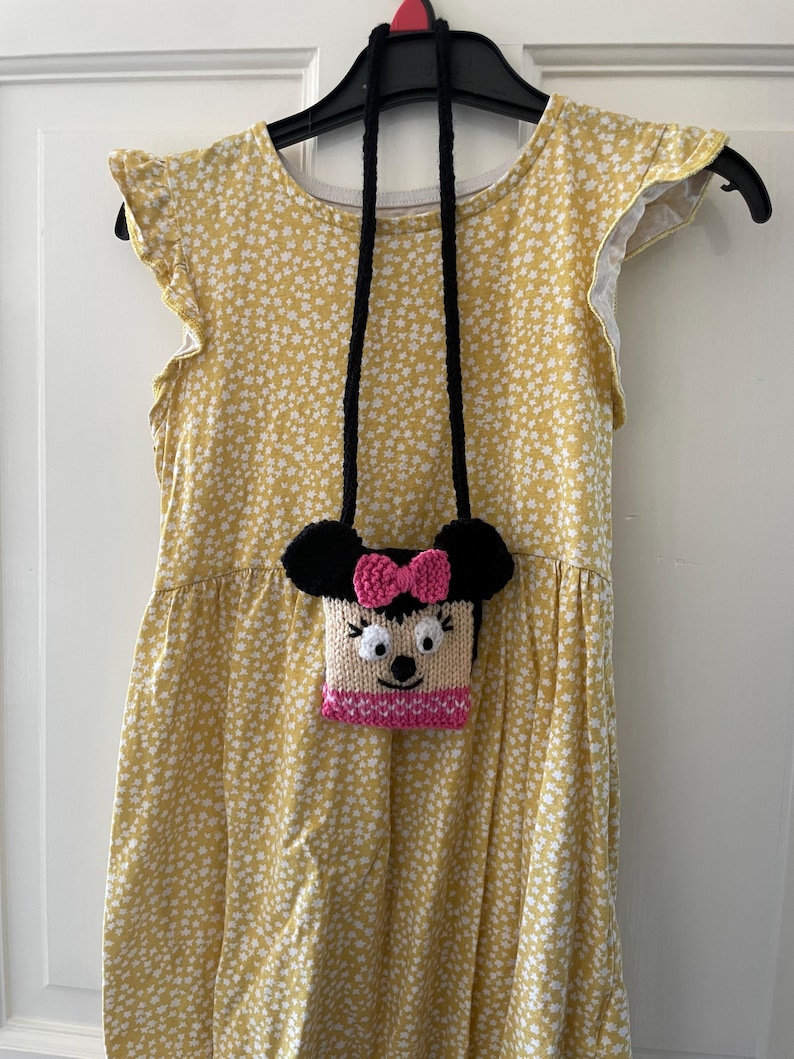 Pu&ograve; includere: Un vestito giallo con stampa floreale bianca e una borsa di maglia nera e rosa con un viso di cartone animato appeso alla tracolla.