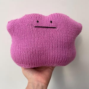 May include: A pink knitted plush toy shaped like a Pokemon character, with black embroidered eyes and a mouth.