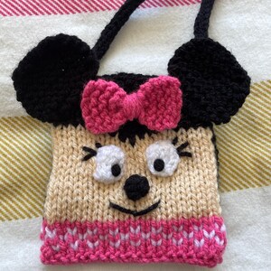 Pu&ograve; includere: Una borsa a mano in maglia a tema Minnie Mouse con un fiocco rosa, orecchie nere e un viso beige con occhi bianchi e un naso nero.