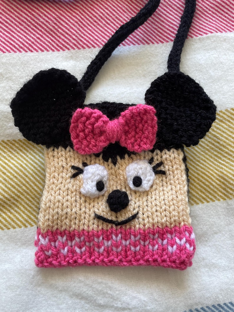 Pu&ograve; includere: Una borsa a mano in maglia a tema Minnie Mouse con un fiocco rosa, orecchie nere e un viso beige con occhi bianchi e un naso nero.