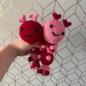 May include: Two handmade knitted dolls in shades of red and pink. Each doll has a round head with black eyes and a smiling mouth. The dolls have heart-shaped antennae and are connected at the body.