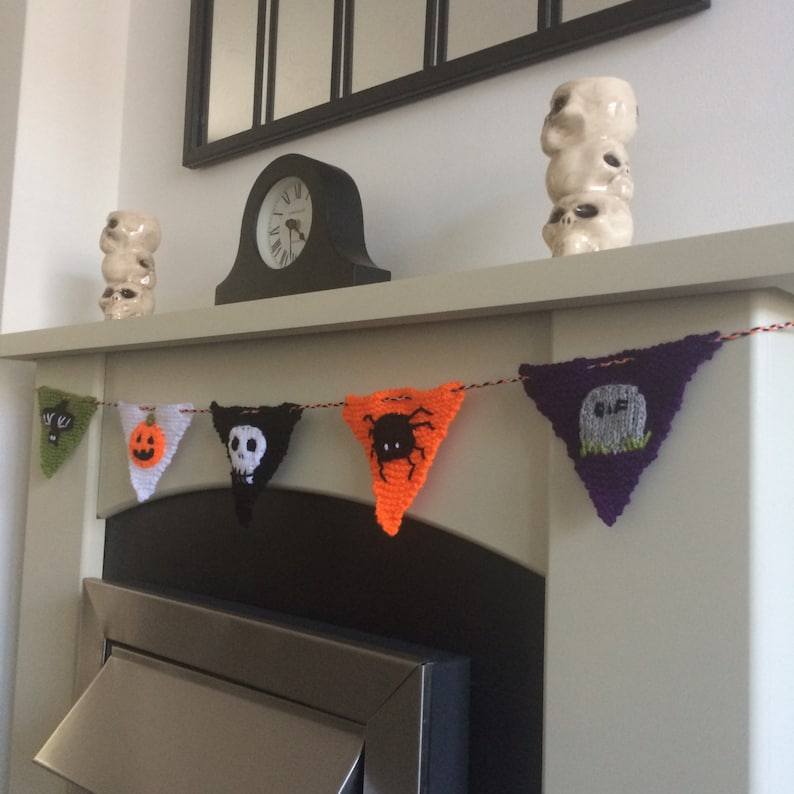 May include: A Halloween banner with knitted triangular flags featuring a pumpkin, a skull, a spider, a ghost, and a bat. The banner is strung across a fireplace mantel.