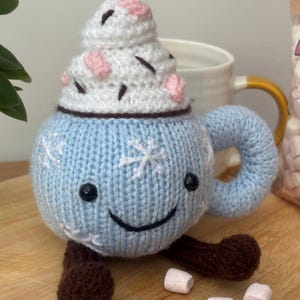 Könnte beinhalten: Eine handgestrickte blaue Tasse mit einem lächelnden Gesicht, Schneeflocken-Details und braunen Füßen. Die Tasse ist mit weißer Schlagsahne bedeckt, die mit rosa Akzenten und Schokostreuseln verziert ist. Ein charmantes, skurriles und gemütliches Dekorationsstück.